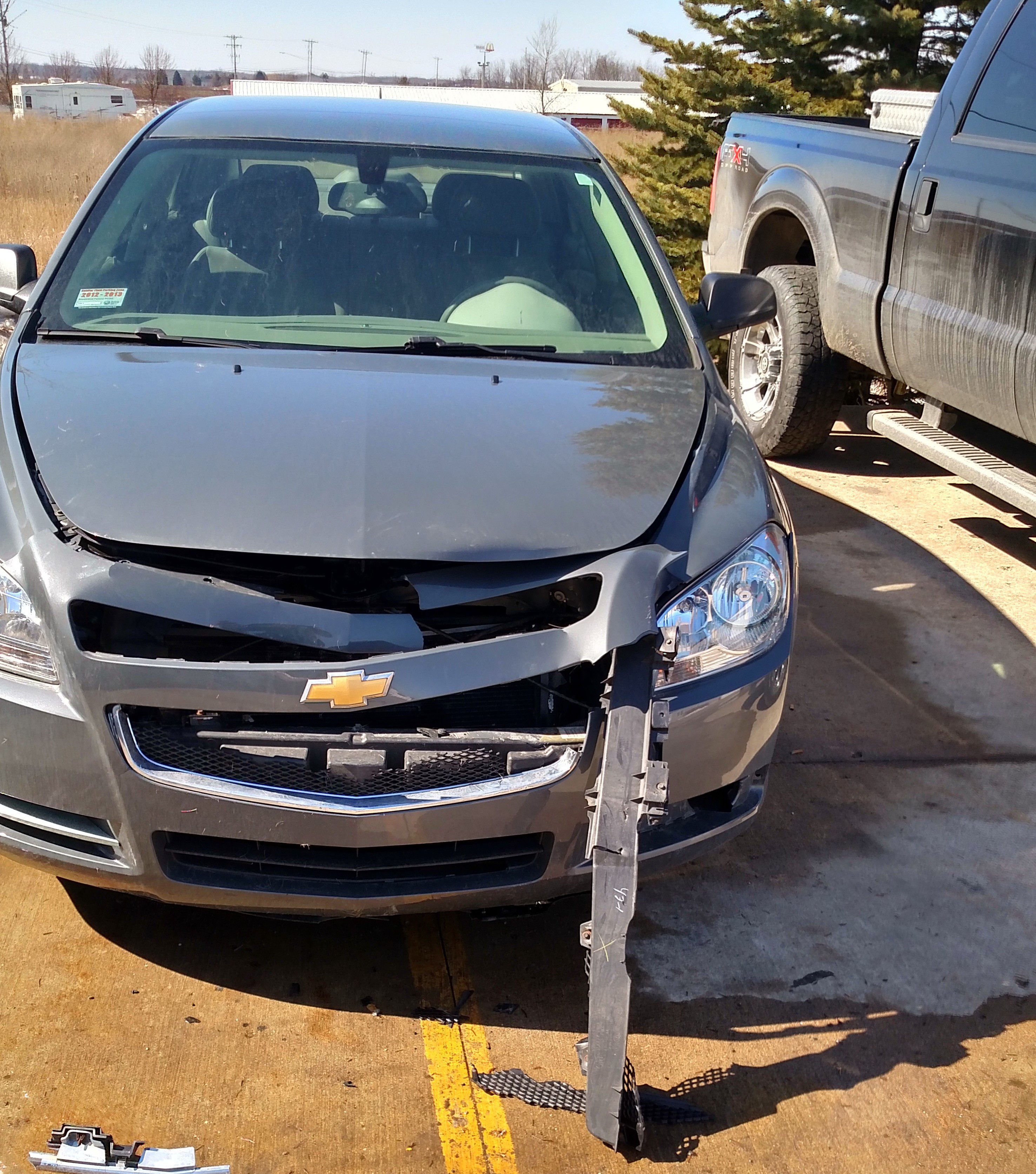 Damaged gray Chevrolet sedan with a detached front bumper and visible front-end collision damage, parked in a vehicle repair facility, illustrating the repair process at Lyle's Collision in Grand Ledge, MI.